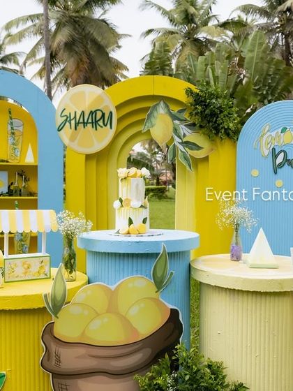 A wide shot of the lemonade party setup, showing the main backdrop, dessert tables, and a "Squeeze the Day" cart on a lush green lawn.