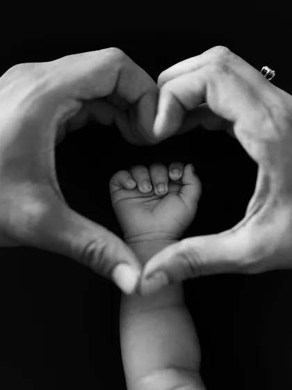 A black and white version of the "heart hands" pose around a baby's fist, a timeless and artistic way to announce your new arrival.