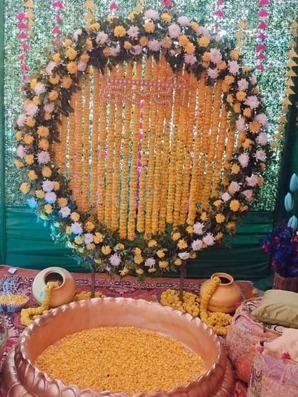 A beautiful Haldi setup with a large brass urli filled with marigold petals. The backdrop is a ring of marigolds against green drapes, with low gadda seating on the side.
