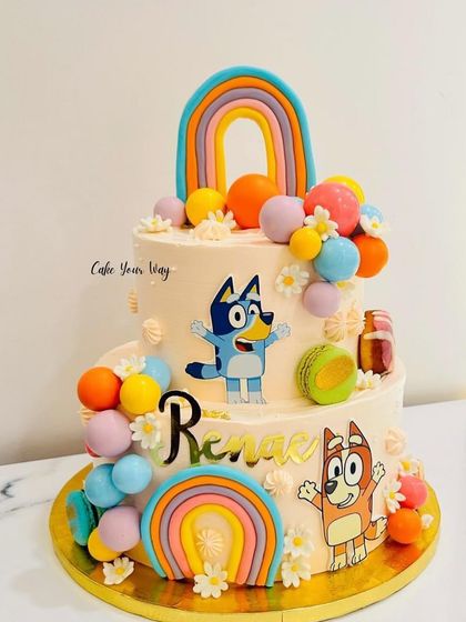 A colorful two-tier cake featuring the popular characters from Bluey, complete with rainbows and macarons.