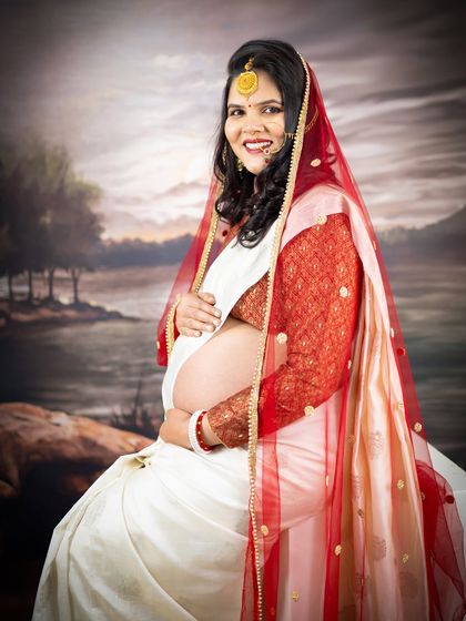 A serene portrait of a mom-to-be in a white and red saree, seated against a painted backdrop. The image has a timeless, painting-like quality.