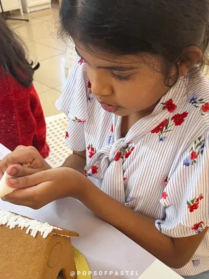 A young girl carefully pipes icing onto the roof of her gingerbread house, concentrating on her design.