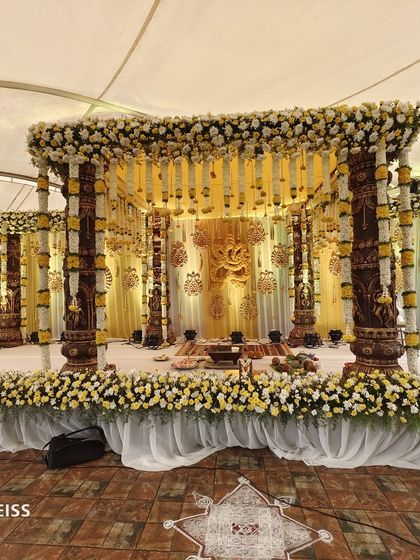 A wide view of an ornate, traditional wedding mandap featuring carved pillars and a ceiling of hanging jasmine and marigold flowers.