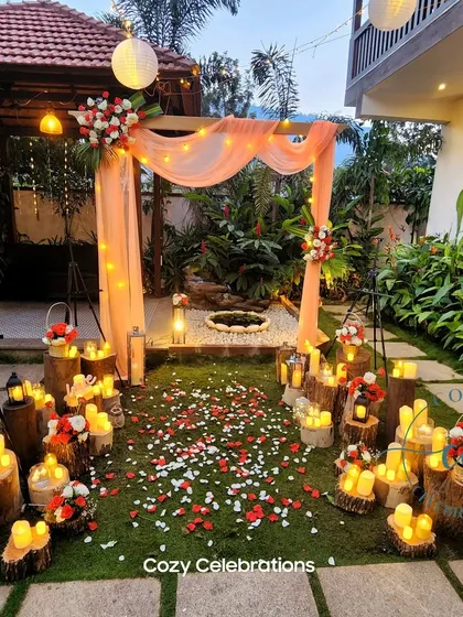 A rustic and romantic garden setup perfect for an anniversary or proposal. A wooden arch draped in peach fabric is surrounded by dozens of LED candles on tree stumps, with a path of scattered flower petals.
