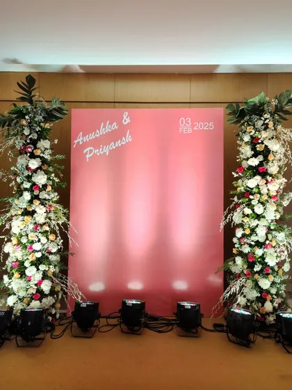 A clean and contemporary photo backdrop designed for a modern couple. The minimalist pink panel with personalized names is flanked by two towering pillars of white and blush florals, offering a chic and stylish spot for pictures.