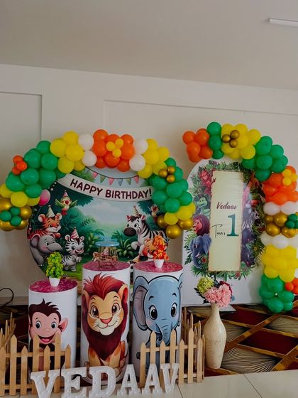 A classic jungle book style setup for a first birthday. It includes a circular backdrop with cartoon animals, character-themed pedestals, and a colorful balloon arch.