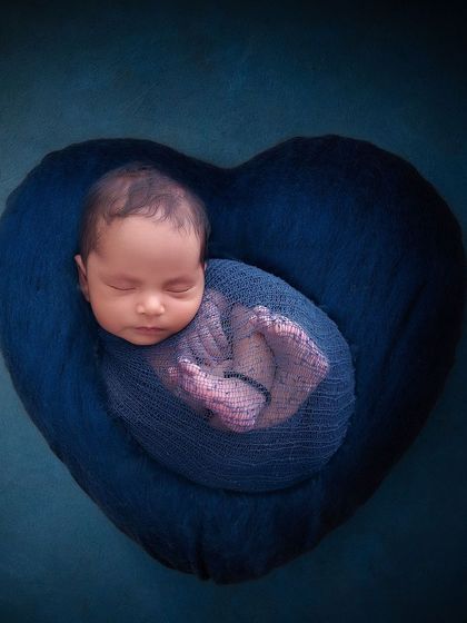 A beautifully swaddled newborn is curled up in the center of a plush, heart-shaped blue pillow, symbolizing a heart full of love.