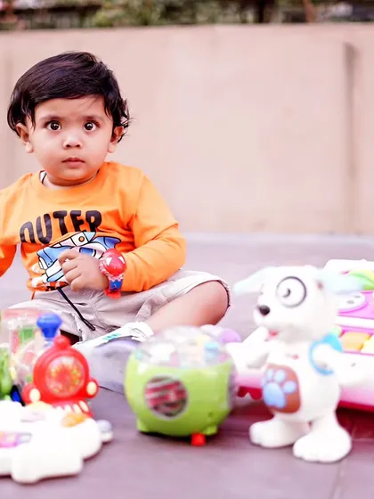 A toddler sits on a patio surrounded by his favorite toys, looking directly at the camera for a curious and engaging portrait.