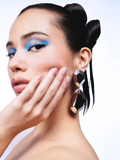 A close-up beauty shot focusing on a pair of large, geometric statement earrings. The model's hand gently touching her face draws the eye upwards, creating a soft and elegant portrait.