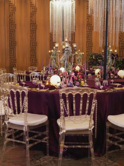 A tablescape that is pure drama. A deep purple tablecloth provides a rich base for gold charger plates, glowing orbs, and a towering candelabra centerpiece, all under the sparkle of a crystal chandelier.