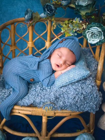 A sleeping newborn in a blue knit outfit, resting on a miniature wicker chair with a candle prop nearby for a serene feel.