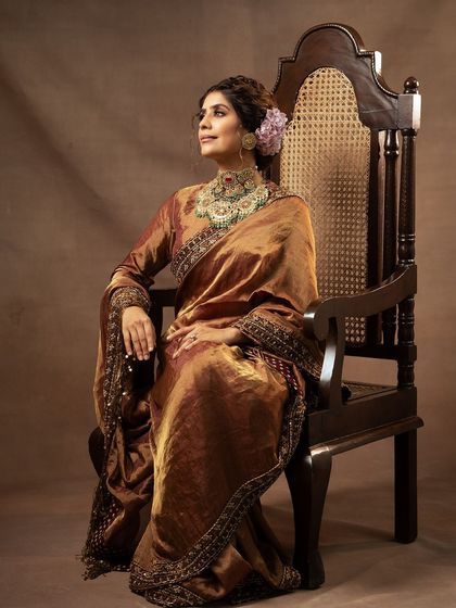 A full-length seated portrait that exudes grace and royalty. The rich, bronze-colored saree and the antique wooden chair create a scene of understated luxury.