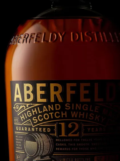 Golden grades. A close-up on the Aberfeldy 12 bottle, focusing on the embossed text and the rich, gold-foiled label against the dark glass. The lighting is designed to pick up every detail.