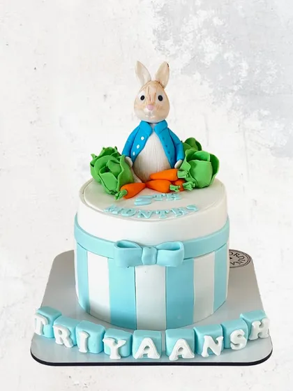 A charming Peter Rabbit cake for Triyaansh. This sweet design features the beloved character on top of a blue and white striped cake, complete with fondant carrots.