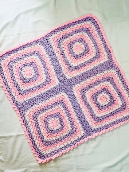A lovely granny square blanket in a classic four-square pattern, using soft pink, purple, and white yarn. The simple scalloped border adds a sweet, delicate finish.