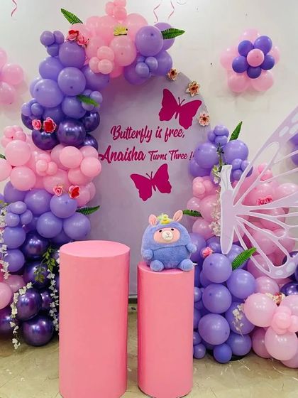 A beautiful butterfly-themed decoration for a 3rd birthday. This setup features a custom backdrop and a balloon arch in shades of purple and pink, with a large butterfly wing prop for photos.