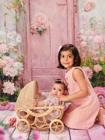 A dreamy sibling portrait with a floral pink backdrop. The older sister proudly stands by her baby sister in a rustic pram, like a little mother.