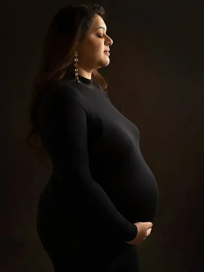 A quiet moment of reflection. This side profile portrait in a form-fitting black dress beautifully highlights the curve of the baby bump against a dark, moody backdrop.