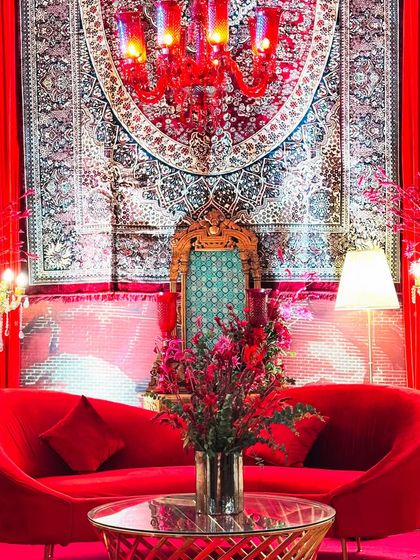 A regal seating arrangement featuring a high-backed throne-like chair against a backdrop of a traditional Persian rug. The entire scene is bathed in red, from the velvet sofa and chandelier to the floral arrangements, creating an opulent and dramatic look.
