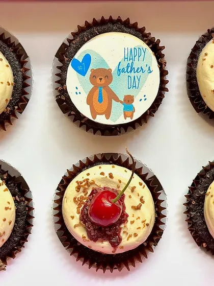 A top-down view of our special Father's Day Black Forest cupcakes. Each one sits on a moist chocolate base and is topped with vanilla buttercream, our homemade cherry compote, chocolate shavings, and a fresh cherry. One cupcake features an edible "Happy Father's Day" topper.