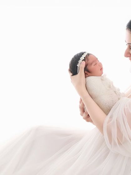 A mother dressed in a flowing white gown smiles at her newborn in a light and airy setting, creating an angelic and ethereal family portrait.