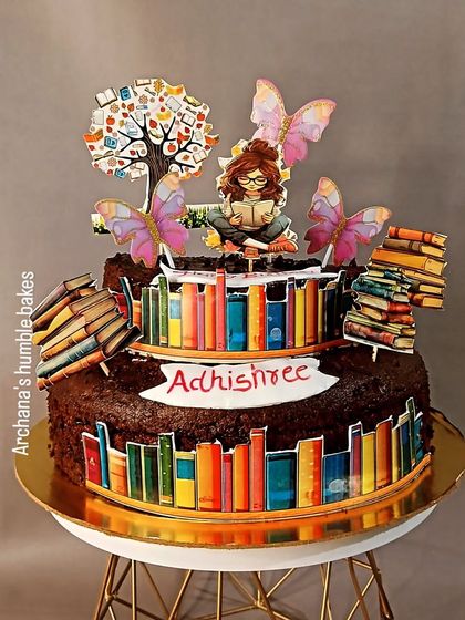 A two-tiered chocolate cake for a book lover, decorated with edible images of bookshelves and a person reading under a tree.