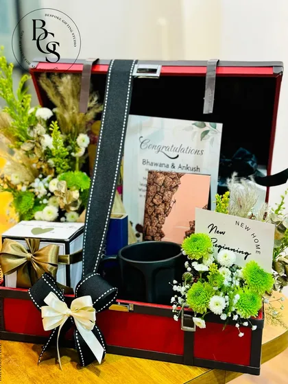 A housewarming or anniversary gift presented in a stylish red and black trunk. It's filled with fresh green and white flowers, a personalized card, and other home-centric gifts to celebrate a new beginning.