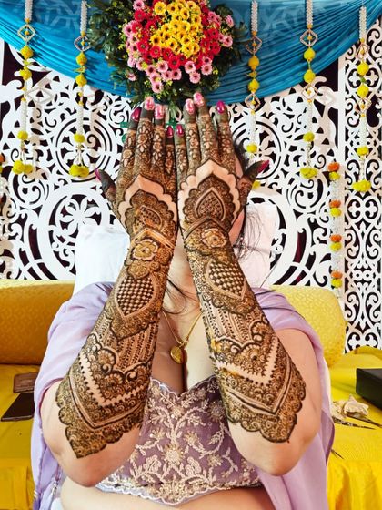 A classic pose showing the full detail of the engagement mehendi, with its heart motifs and checkerboard patterns, against a festive backdrop.