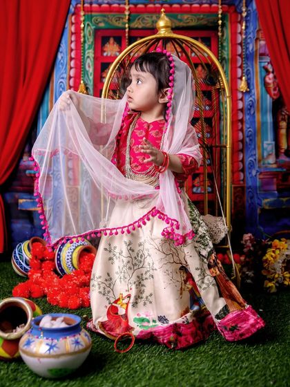 A full-length shot of a little girl in a beautiful Radha costume, complete with a sheer dupatta. The colorful setup with red drapes and floral decorations creates a rich, festive atmosphere.