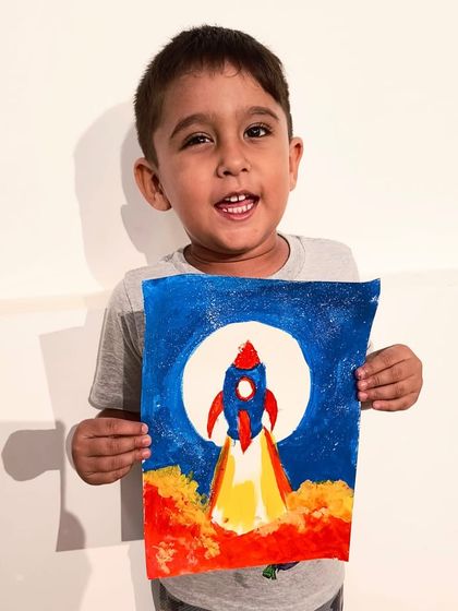 A very proud young artist holds up his dynamic painting of a rocket blasting off to the moon.
