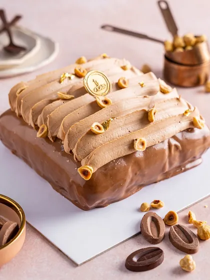 Our La Noisette cake, a square-shaped travel cake celebrating the rich flavour of hazelnut. A glossy chocolate glaze covers the cake, which is topped with a piped gianduja mousse and toasted hazelnuts.