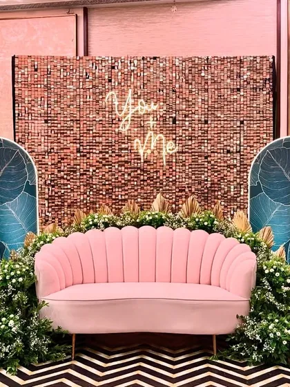 This Sangeet stage combines bohemian and tropical elements. The pink scalloped sofa is set against a backdrop of a shimmer wall and panels with large monstera leaf prints.