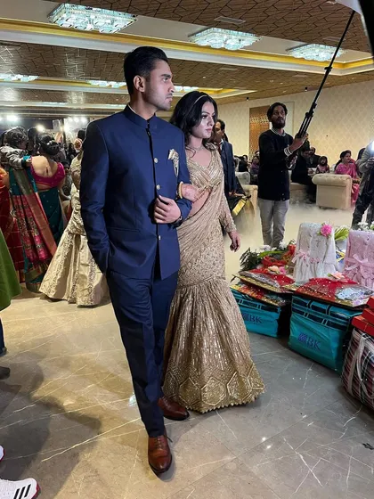 A candid shot of a couple making their entrance at a wedding. The groom is in a classic navy bandhgala, and the bride is in a stunning gold fish-cut lehenga saree.
