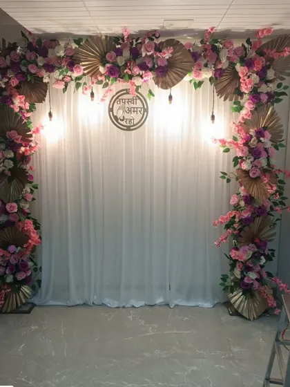 A floral arch in pink and purple for a home-based Jain ceremony. The design includes golden palm leaves and a central metallic sign with "Tapasvi Amar Raho".
