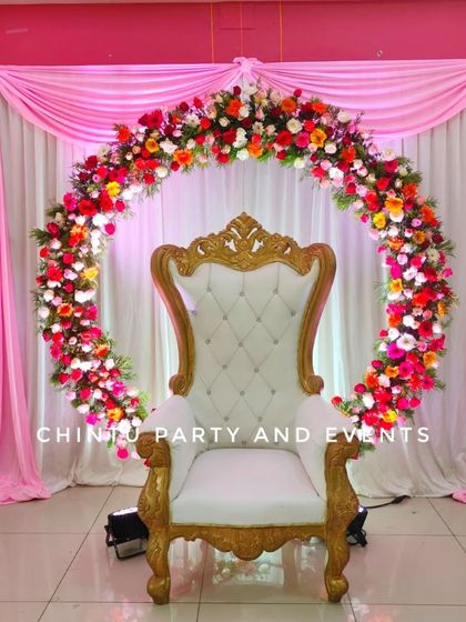 A beautiful Half Saree function backdrop featuring a circular floral arch. The setup includes a white throne chair against a pink and white draped background.