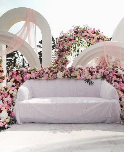 A dreamy pastel pink floral stage for a daytime event. The cascading flowers over the sofa and the modern white arches create a soft, romantic, and incredibly photogenic setting for the couple.