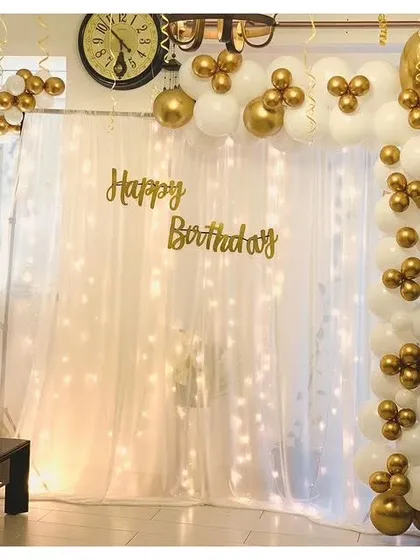 A simple yet elegant birthday decoration with a white and gold balloon garland, sheer curtain, and fairy lights.