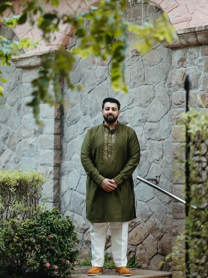 A classic portrait of the groom in his olive green kurta. The stone wall background and natural framing from the leaves create a rustic and handsome look.
