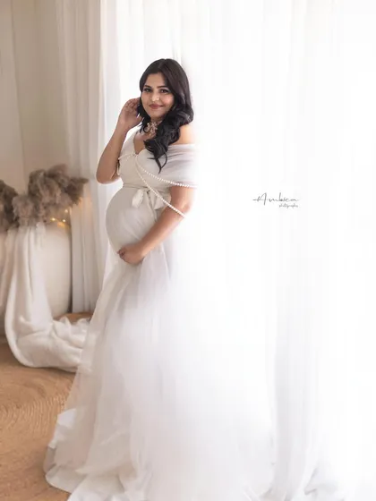 A full-length portrait showcasing the beautiful white gown against the bright, airy backdrop. Her happy expression and relaxed pose make for a lovely, natural maternity photo.