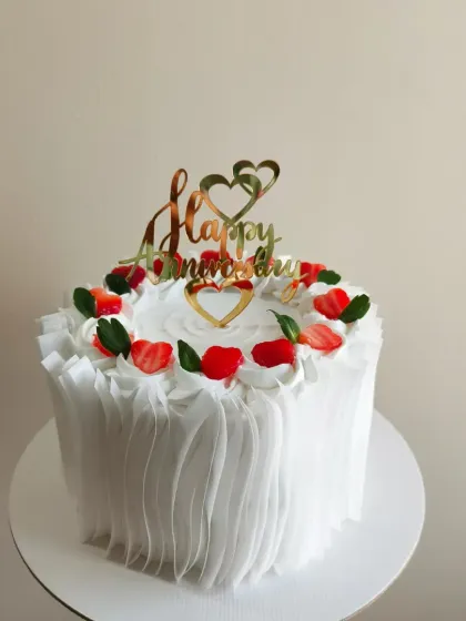 A beautiful white anniversary cake decorated with a crown of fresh, heart-shaped strawberries and a golden "Happy Anniversary" topper. The ruffled frosting on the side adds an elegant touch.