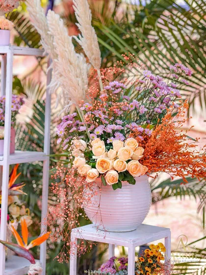 A close-up of a lush floral arrangement in a white ceramic vase, with soft peach roses and fluffy pampas grass.