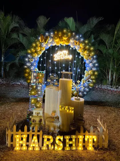 My decorations are not just for indoors. I can create stunning backdrops for outdoor and garden parties, like this setup for a birthday celebration. The circular balloon arch with chrome gold and silver balloons stands out beautifully against the natural greenery, especially at night with the neon sign and marquee letters lit up.