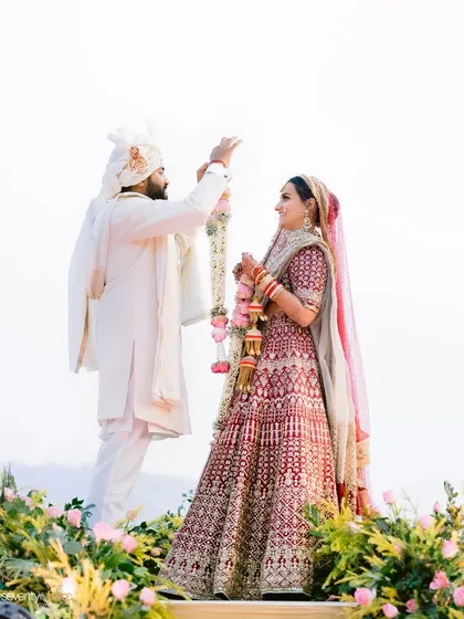 The moment of the varmala exchange, set against the sky and a line of pink flowers, a simple yet beautiful composition.