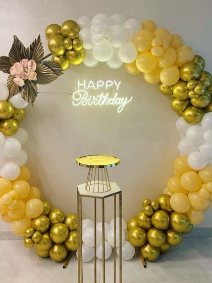 A chic birthday decoration with a circular balloon arch in yellow, white, and gold. The setup includes a cake stand and a 'Happy Birthday' neon sign.