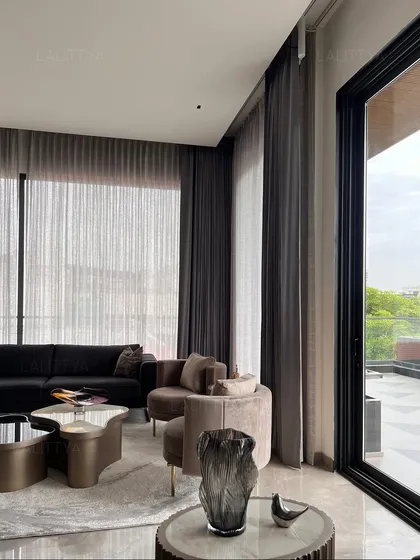 Natural light floods this modern living area through floor-to-ceiling windows, highlighting the soft, curved forms of the armchairs and coffee table. The sheer and heavy curtains provide options for light control while maintaining a sophisticated look.