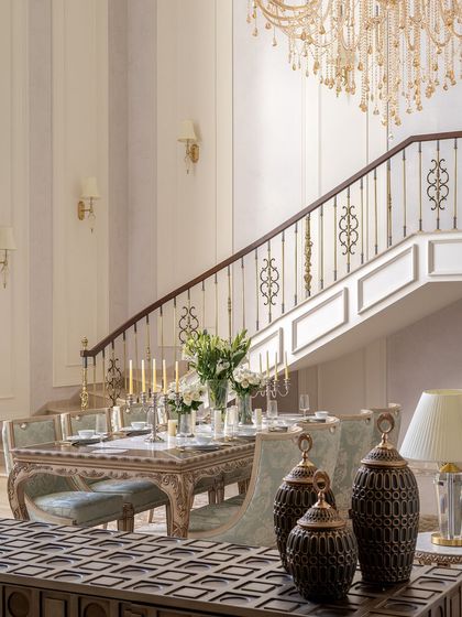 The grand staircase of The Ivory Royale serves as a stunning backdrop to the formal dining area. The combination of the sweeping staircase, crystal chandelier, and elegant table setting creates a scene of timeless sophistication and grandeur.