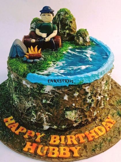 A unique camping-themed cake for a husband's birthday. This creative design features a fondant man by a campfire, a tent, and an edible water effect, all on a rustic, textured cake.