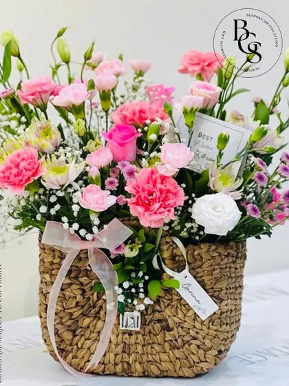 A charming purse-shaped wicker basket filled with a delicate arrangement of pink carnations and white accent flowers. This unique design is a whimsical and stylish way to gift flowers.