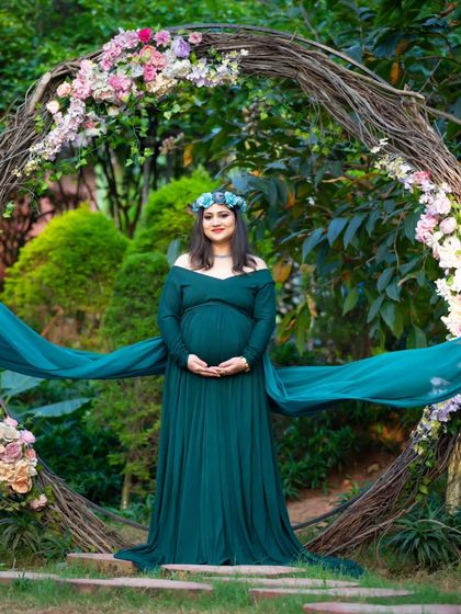 A majestic outdoor portrait. The mom-to-be stands in a deep green gown before a large rustic wreath adorned with flowers, creating a powerful and earthy image.