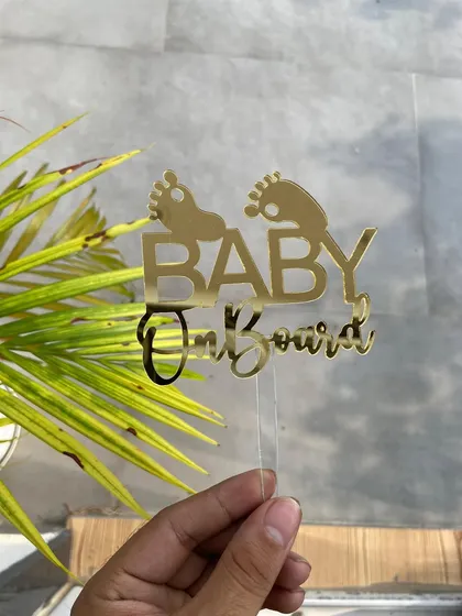 The "Baby on Board" topper held against a natural, green background, highlighting its vibrant gold color.